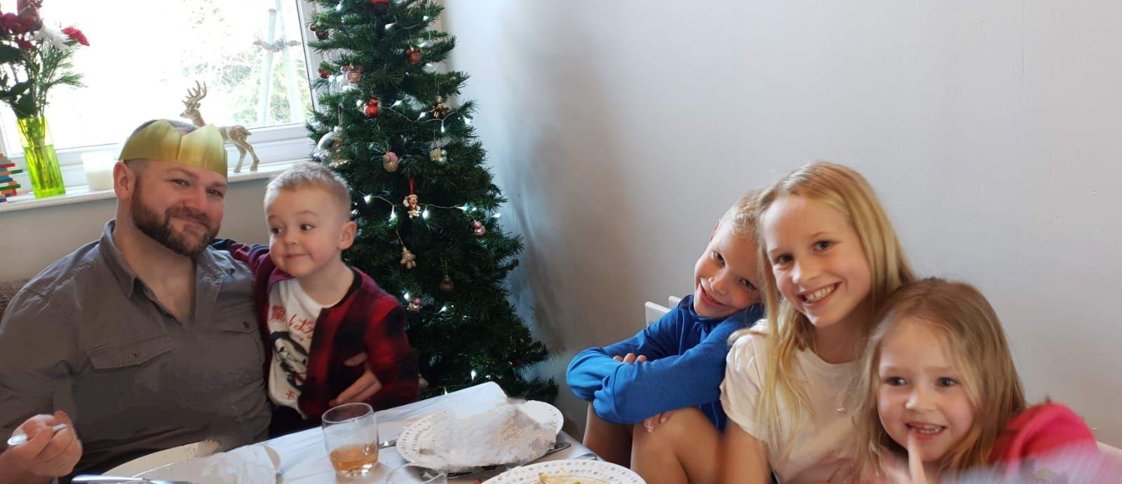 James sitting with four children around a dinner table around Christmas time. A Christmas tree can be seen in the background. All are smiling.