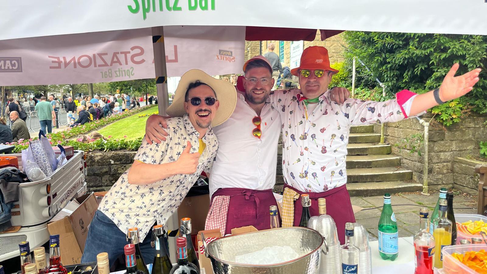 Three men wearing white floral shirts have their arms around each other. In front of them is a table with bottles of spirits next to an ice bucket. Behind them, crowds of people sit on grass verges to watch the Ilkley Cycle Races.