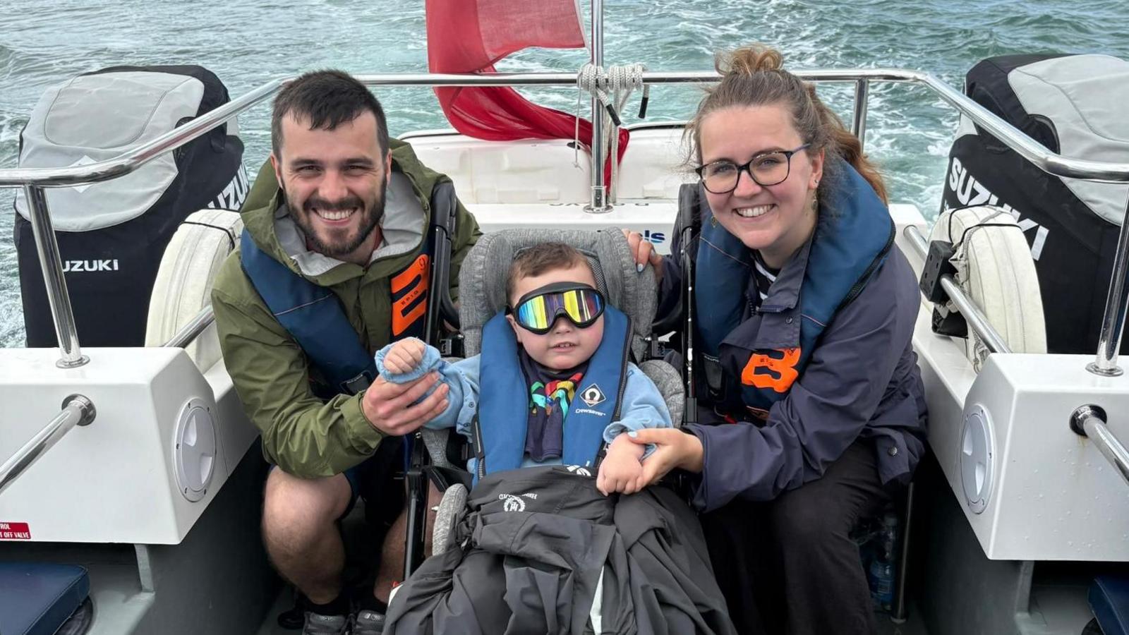Lucy and Joel Capes sit either side of their son at the back of a moving boat with two boat engines either side and the water behind them. The son is wearing tinted goggles. They are all wearing life jackets.