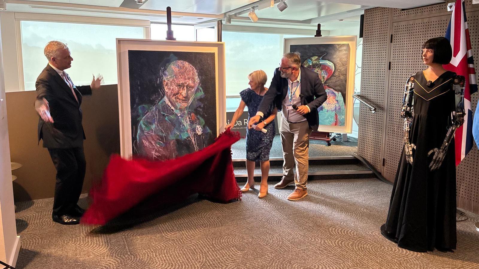 The portrait of King Charles III unveiled at the United Nations' AI for Good Global Summit. Ai-Da is standing to the side while her inventor Aidan Meller and two other speakers pull the red veil down to showcase the portrait. Ai-Da's portrait of the late Queen Elizabeth II is behind them.