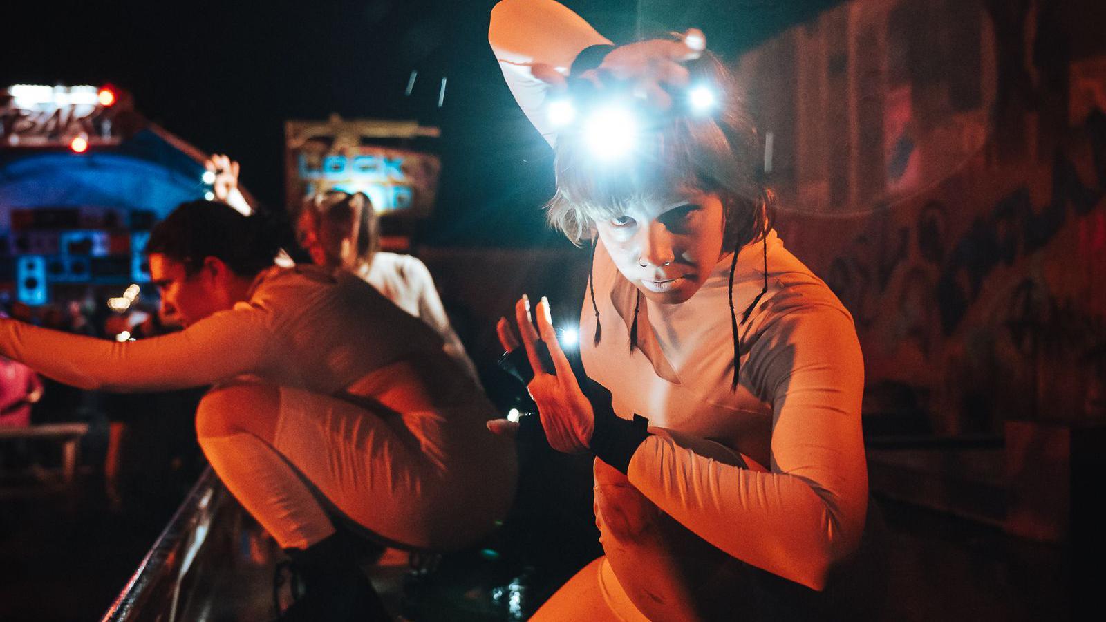 Two stage performers at one of FEAR Scream Park's night time events. One performer is leaning their body towards the camera and has one hand on their head and the other in front of them. They are both wearing white tightly-fitted outfits and are illuminated by the lighting.