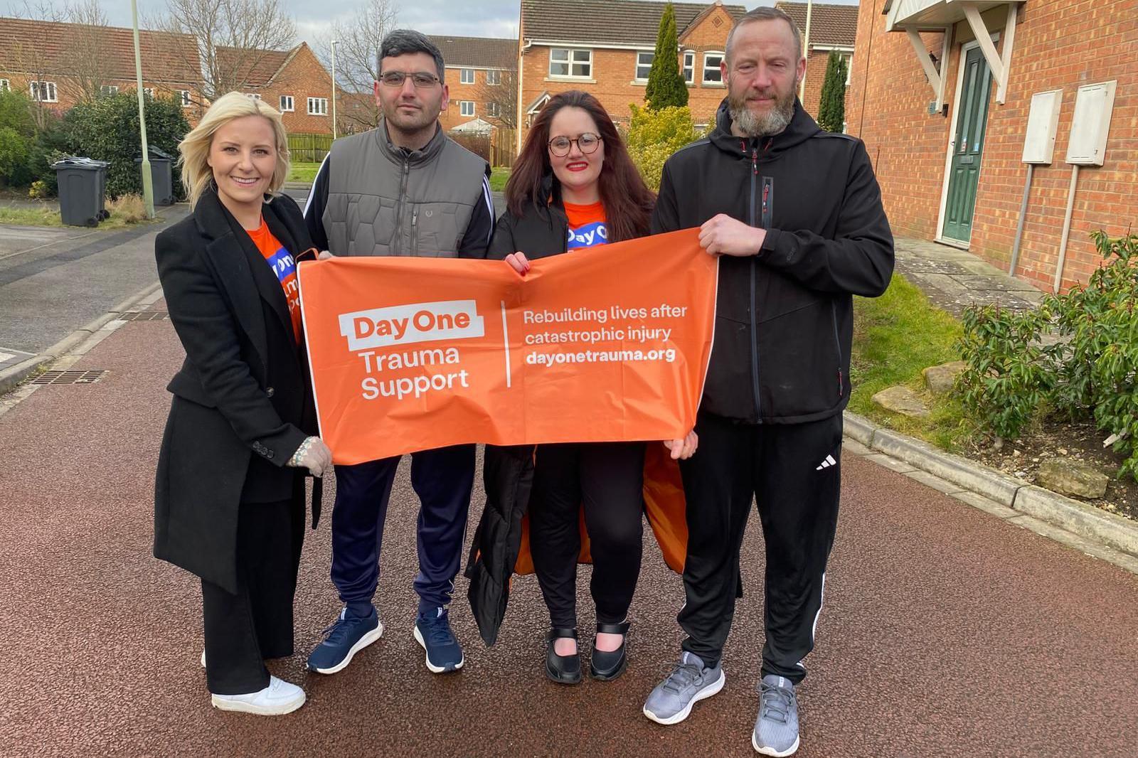 Cat Neill stands with three other people holding up a "Day One Trauma Support" banner. They are on a residential street.