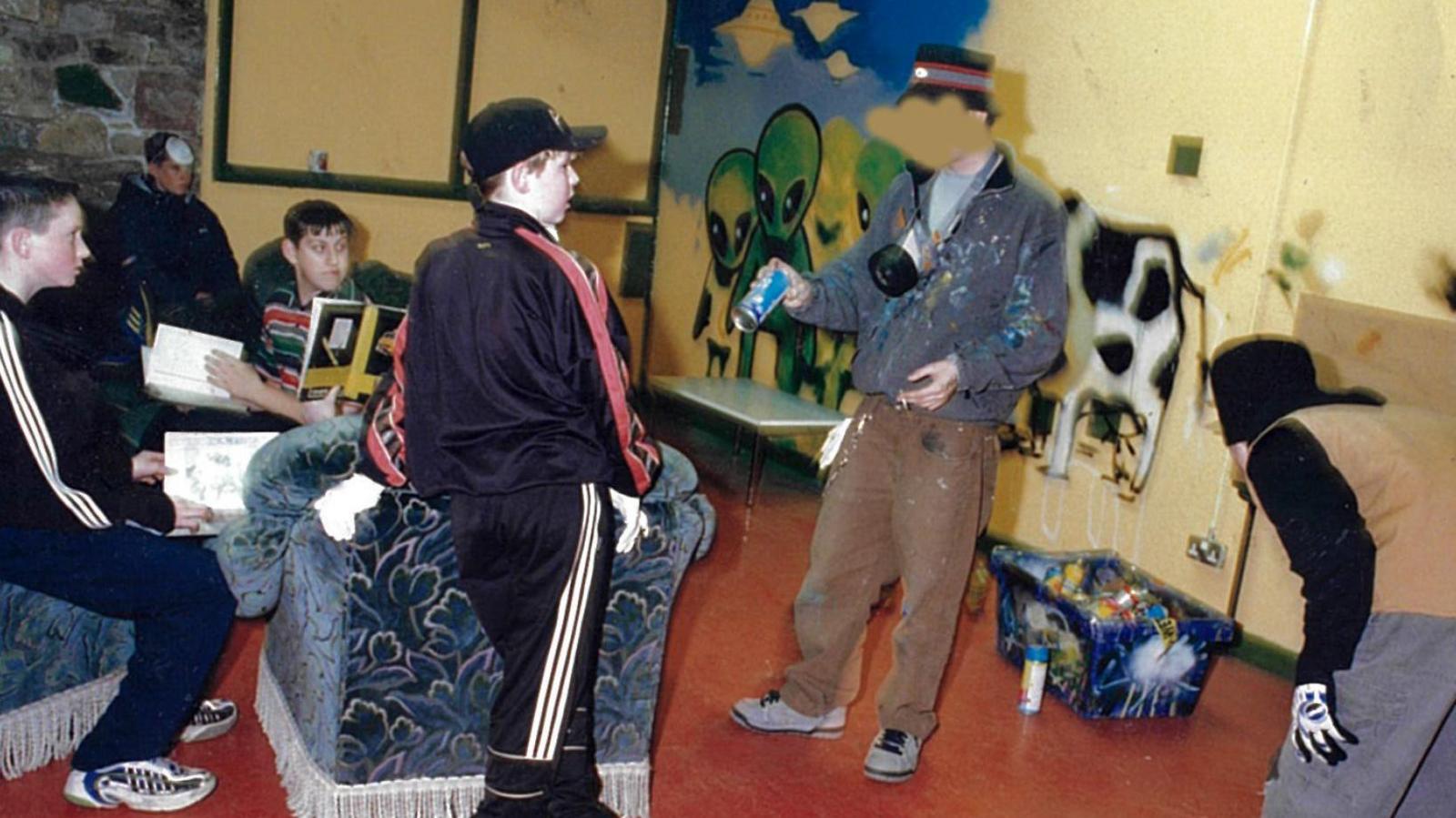 Banksy addresses a room of young people, holding a blue spray can and has a mask hanging from his neck. He is wearing a bucket hat, a paint-covered zip-up top and brown trousers. Behind them is a half-painted mural featuring aliens, spaceships and a cow.