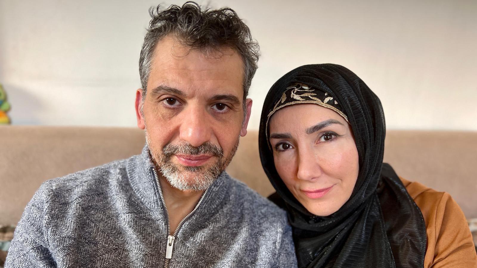 Couple sitting next to each other looking directly at the camera. The man has dark, graying hair and a board. The woman is wearing a black hijab. 