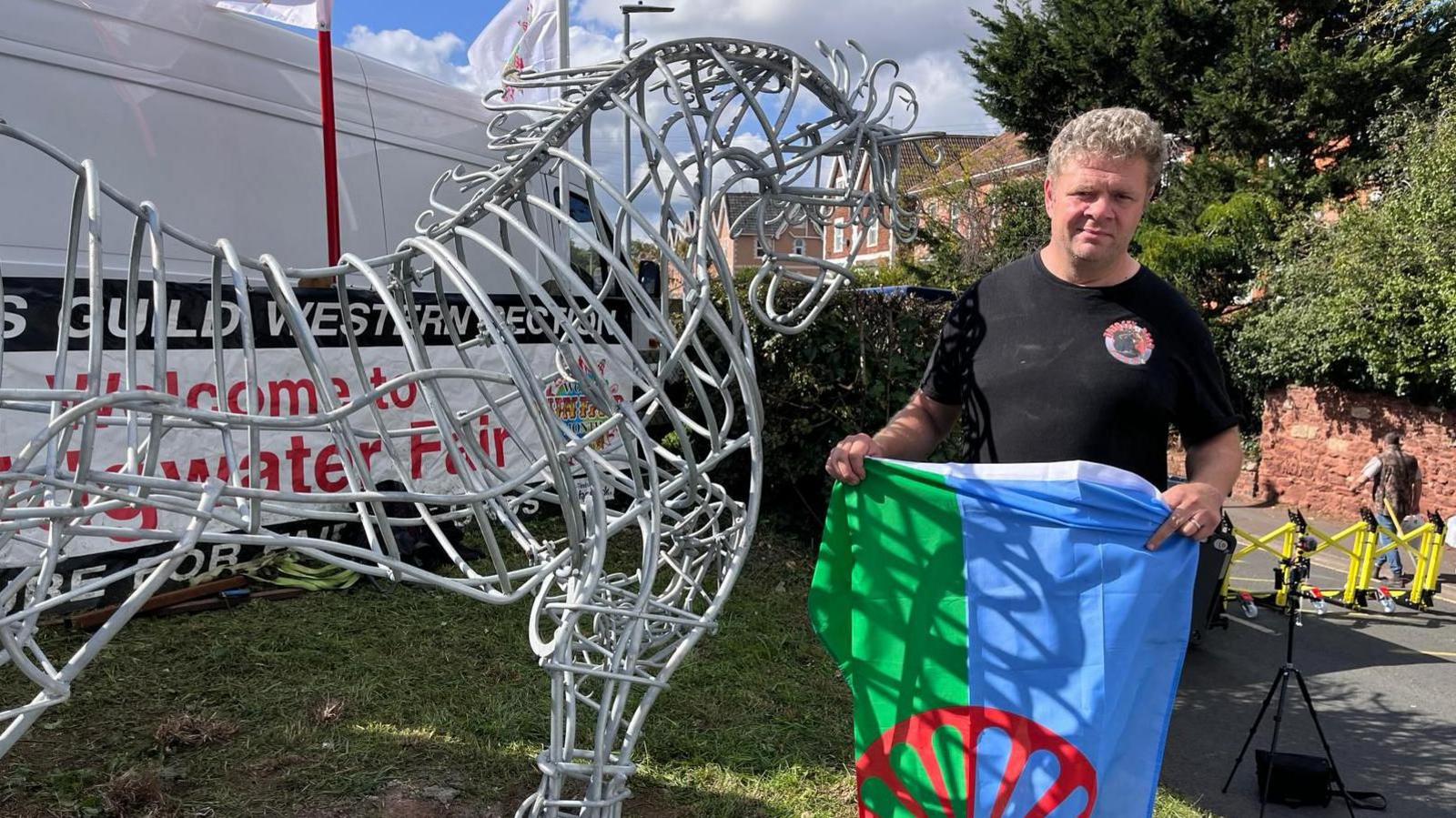 A man is standing next to a sculpture of a horse, wearing a black T-Shirt holding the flag symbolising the Romany community. He is holding the flag so it falls vertically, with the left half as you view it green and the right half blue. A red cart wheel sits in the middle of the flag. The sculpture is made of metal, with various loops of silver-coloured steel connected to each other by rods. The horse's neck is bent, as though it is bending towards the man, It is on a patch of grass and a sign is just visible through it which reads 'Welcome ot Bridgwater Fair'.