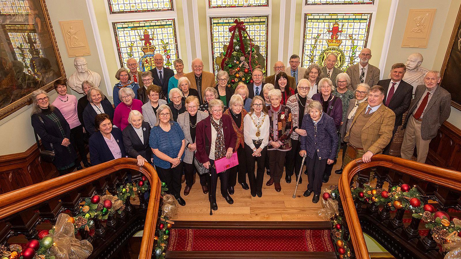Members of the Inverness Field Club gathered on the grand staircase at Inverness Town House. There are stained glass windows and Christmas decorations behind them.