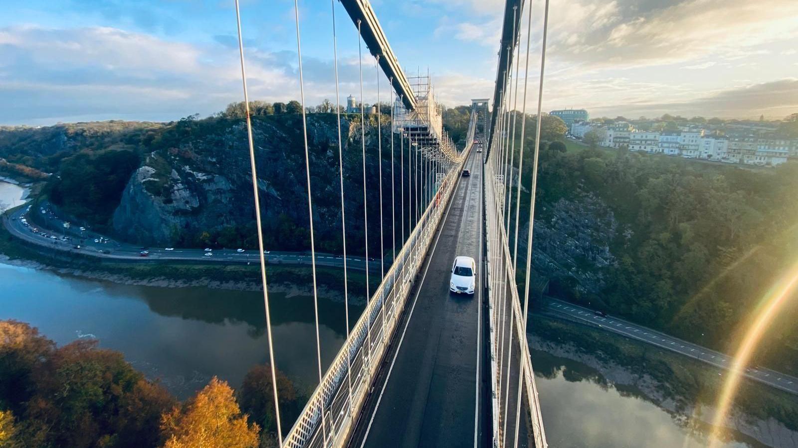 A view of the Clifton Suspension Bridge as taken from high up on the wire suspension cables. It shows the road along the bridge, with a white car driving towards the camera. The sun is setting on the right of the camera and casting a light streak across the corner. 