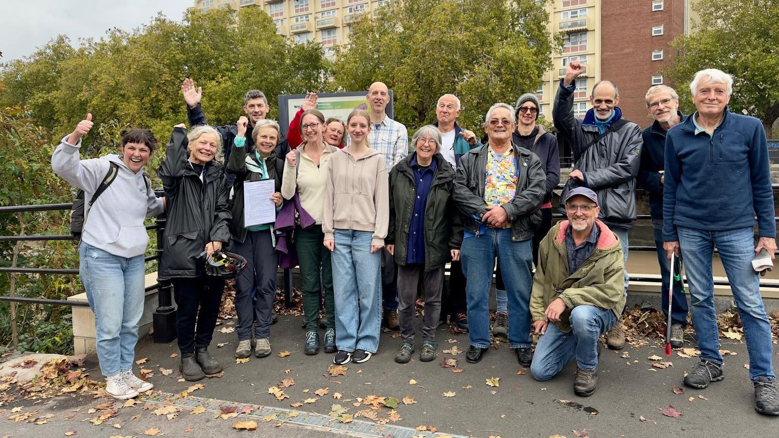 A group of adults in casual clothes stand on a pavement next to the River Avon in the centre of Bristol. They are smiling as they look at the camera. Behind them is a large block of flats with a brown exterior. There are a few autumn leaves scattered on the ground.