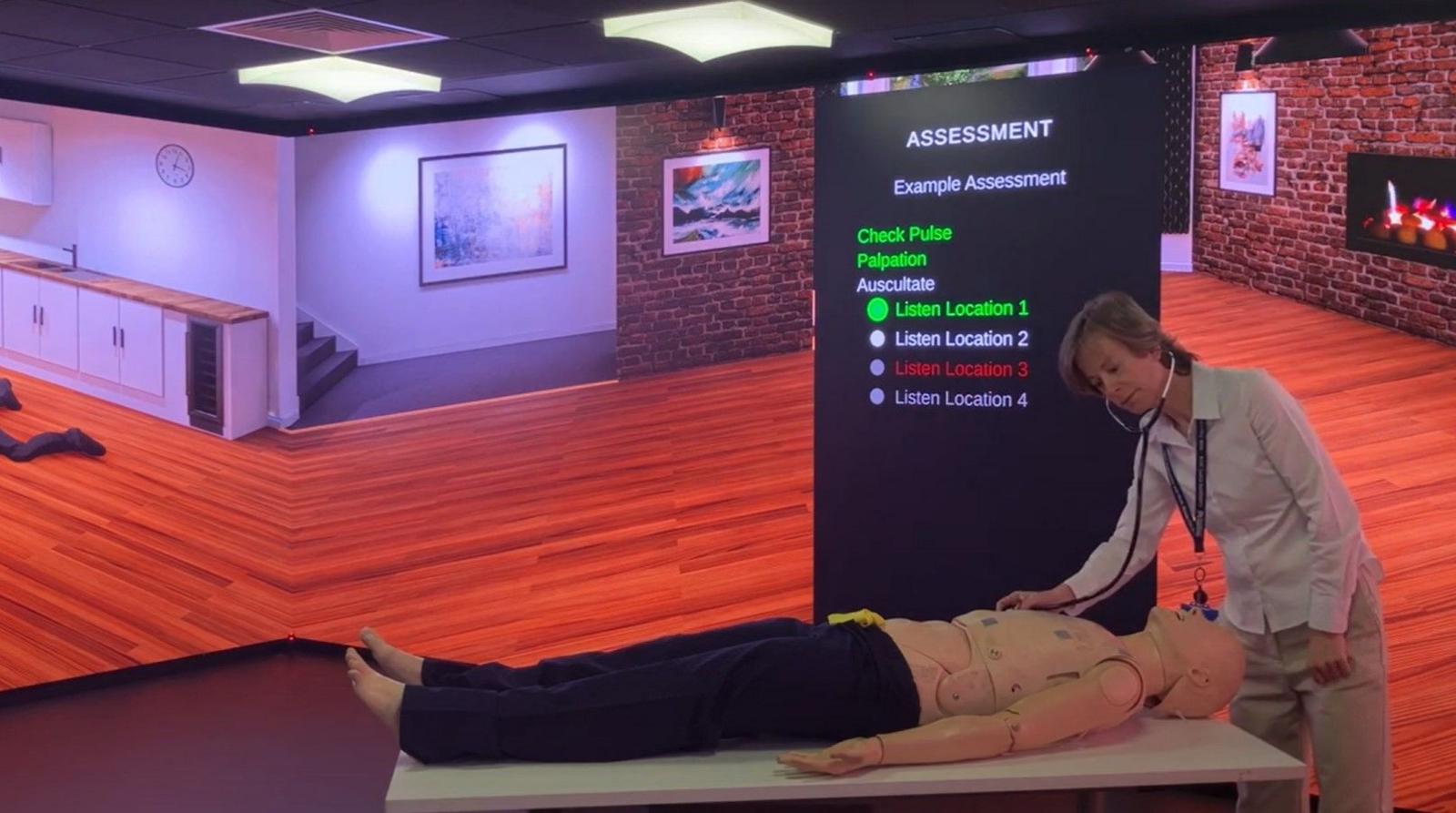 A woman listens to a dummy patient's chest through a stethoscope, surrounded by virtual display of a room in a house, and a virtual screen showing the progress of the examination. 