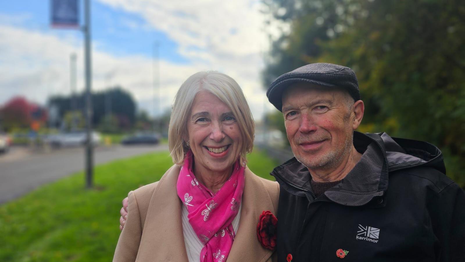 Carolyn Bardill has short blonde hair, she is wearing a beige coat with a large red poppy attached and is wearing a bright pink scarf. Her brother Jeff has his arm around her, he is wearing a black flat cap and a black coat with a small poppy pin on it. The background is blurred but you can see they are standing near a grass verge extto a road.