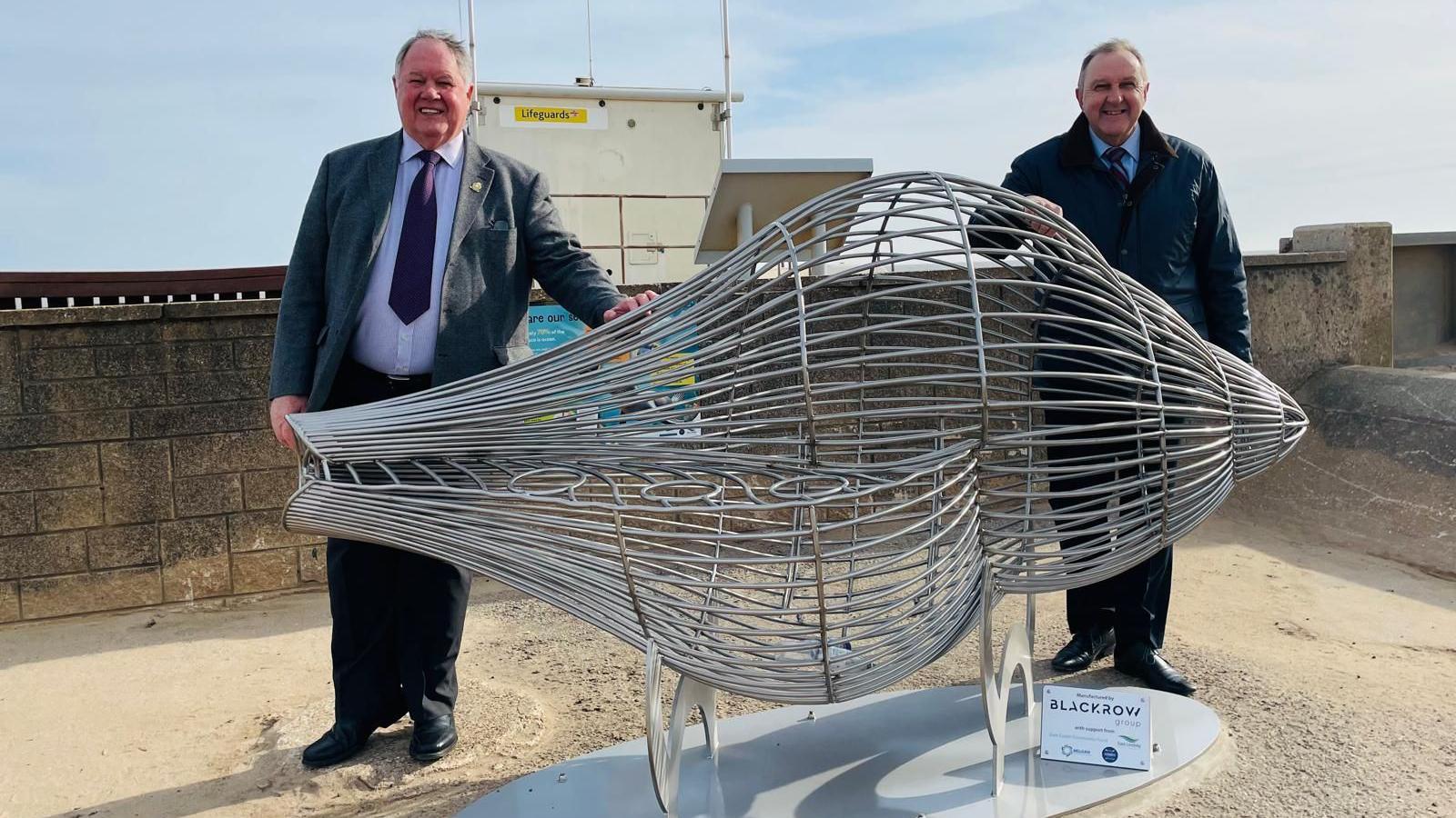 Two men smiling and wearing suits and stood behind a large metal bin in the shape of a seashell.