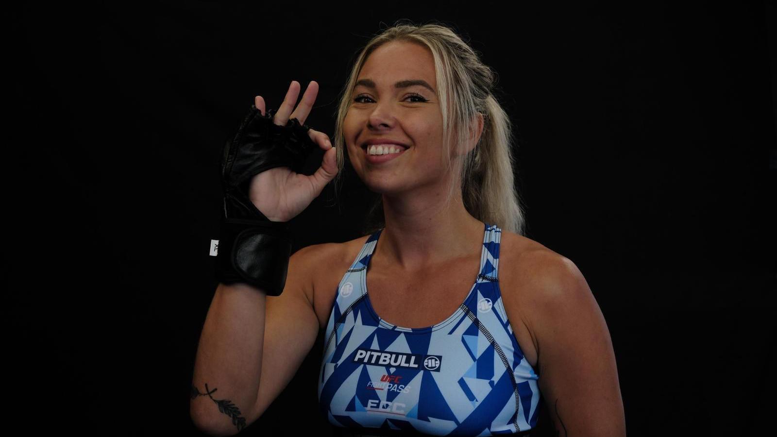 Mia Trevorrow, with her hand making an ok sign. She is wearing a black fingerless glove on her hand. She it wearing a blue sports bra. She is smiling at the camera and her hair is blonde and in a ponytail. The background behind her is black.