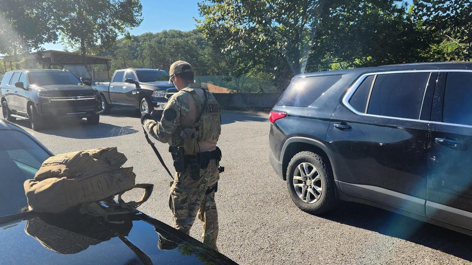 A man in tactical gear holding a gun surrounded by cars on a highway.