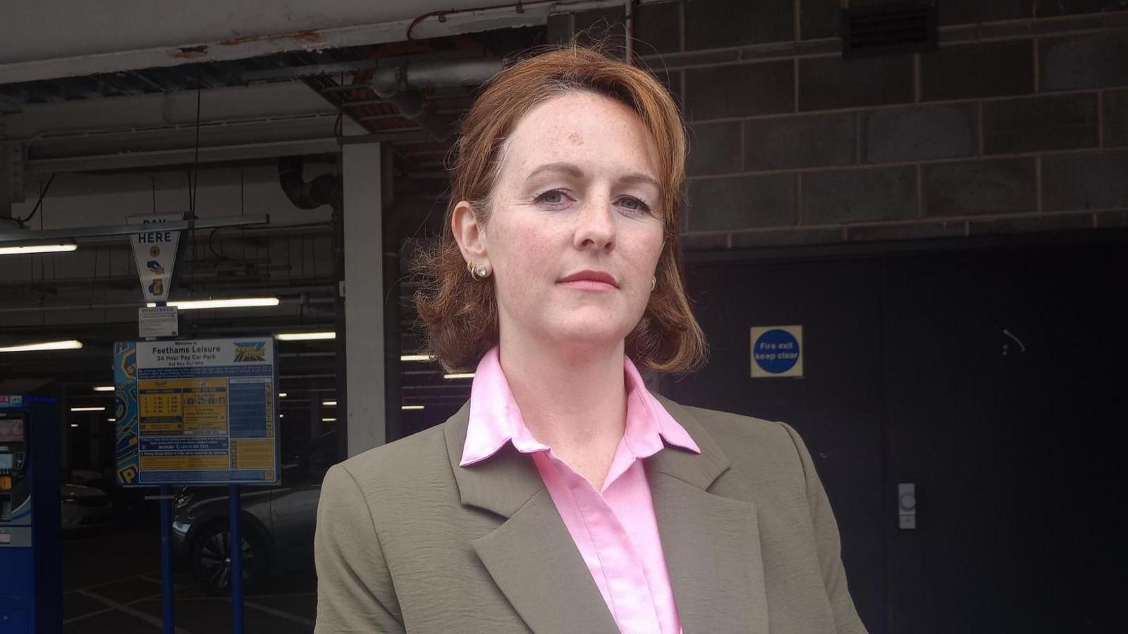 A woman with short red hair wearing a pink shirt and brown jacket stands at the entrance of a covered car park