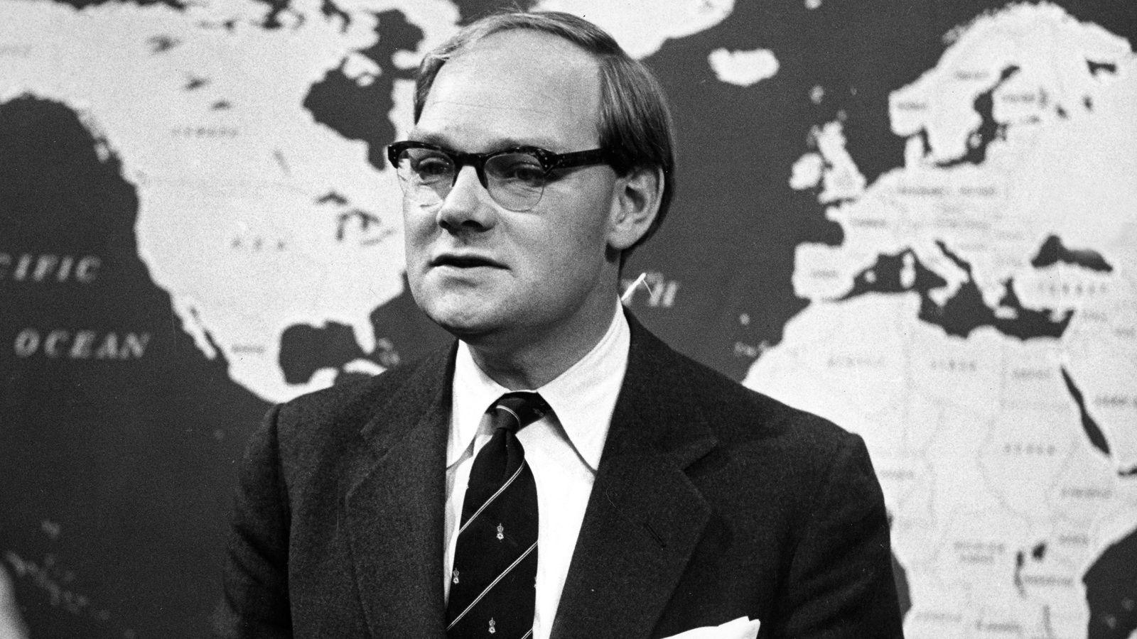 A black and white image showing a man in his 40s with glasses and balding hair, from the chest up, wearing a dark suit jacket, a white shirt and a dark tie with thin slanting stripes and some unidentifiable small emblems between the stripes. He is standing in front of a large map of the world on the wall behind him.
