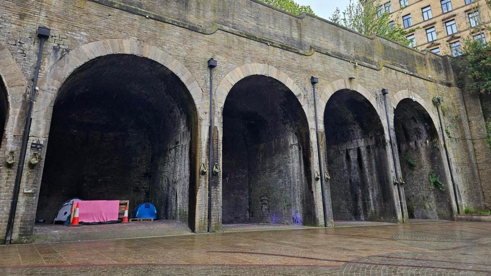 A row of brick railway arches. Under one arch there are tents and traffic cones.