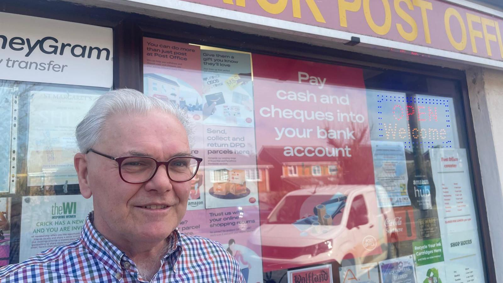 A man with swept back white hair and glasses standing outside a post office with numerous adverts displayed in the windows.