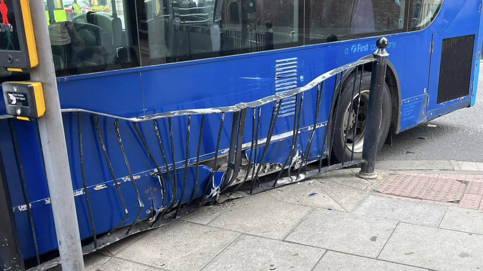 A damaged lower side of a blue bus with a damaged black guardrail in front of it with a pedestrian crossing button in the foreground