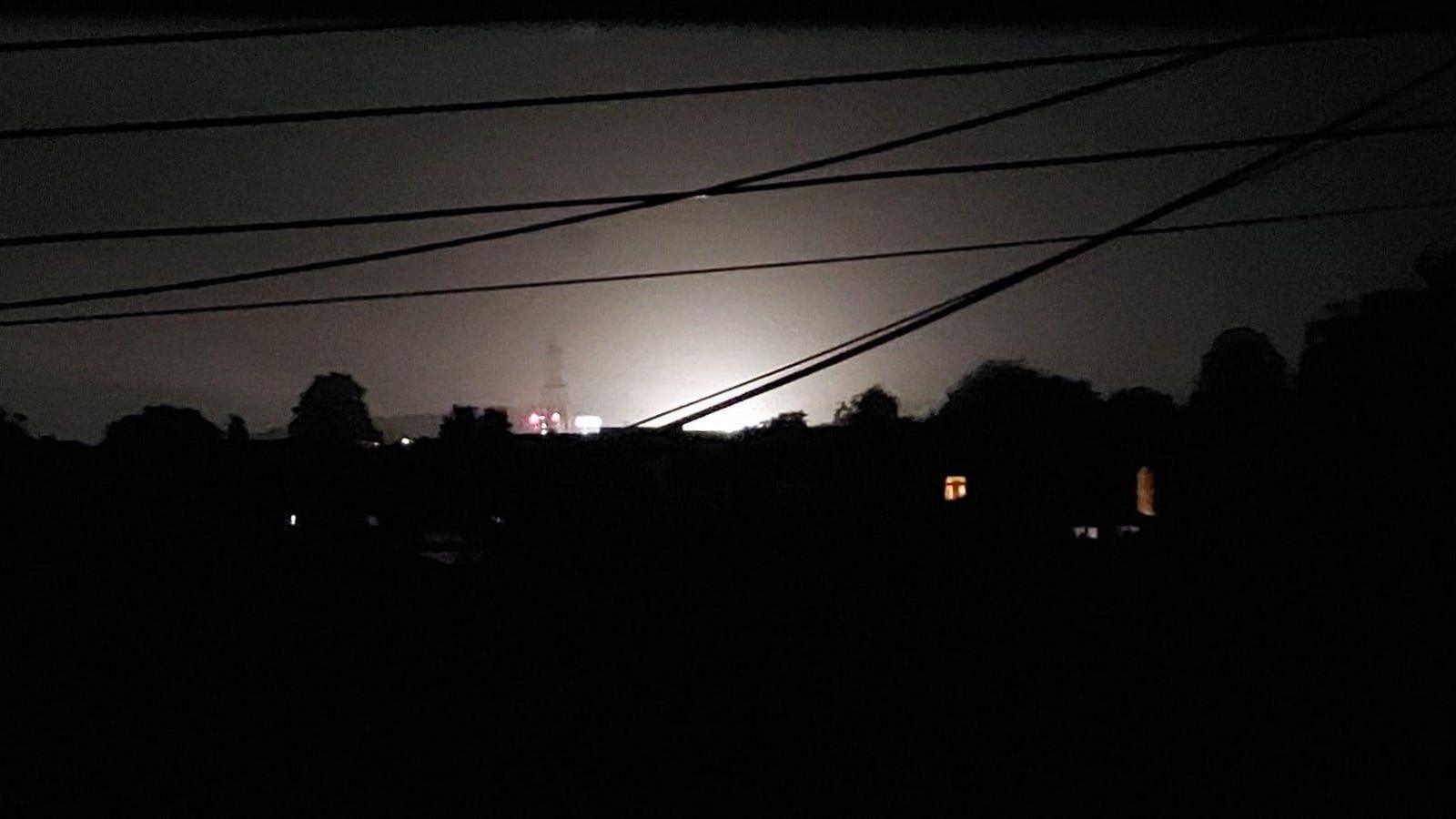 Lights illuminate the sky. In the foreground are silhouettes of trees and electricity wires.