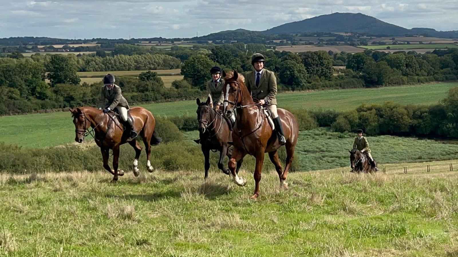 In the foreground are three horses being ridden by hunters wearing tweed jackets, beige jodhpurs, and long black riding boots. The horses are various shades of brown and chestnut - there are more riders and horses in the background. They're riding along a ridge with the Wrekin clearly seen in the background - the weather is sunny and the grass is bright green. 