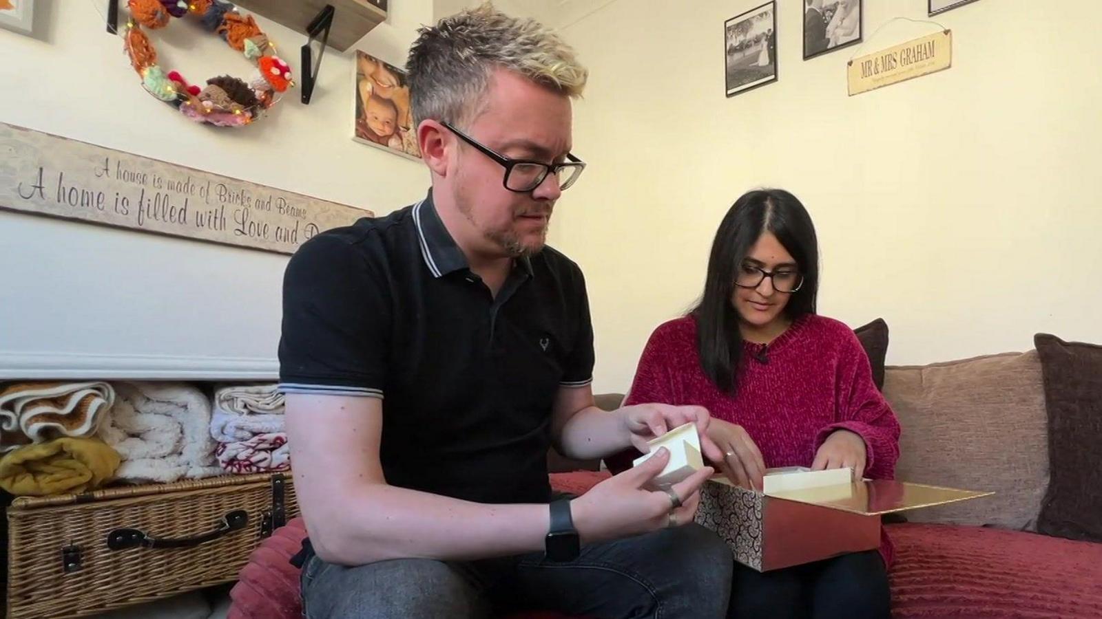 A man and a woman holding a box with small items in them. The man, wearing a black polo shirt, is holding a small box, while the woman, wearing a pink jumper, is holding the box on her lap.