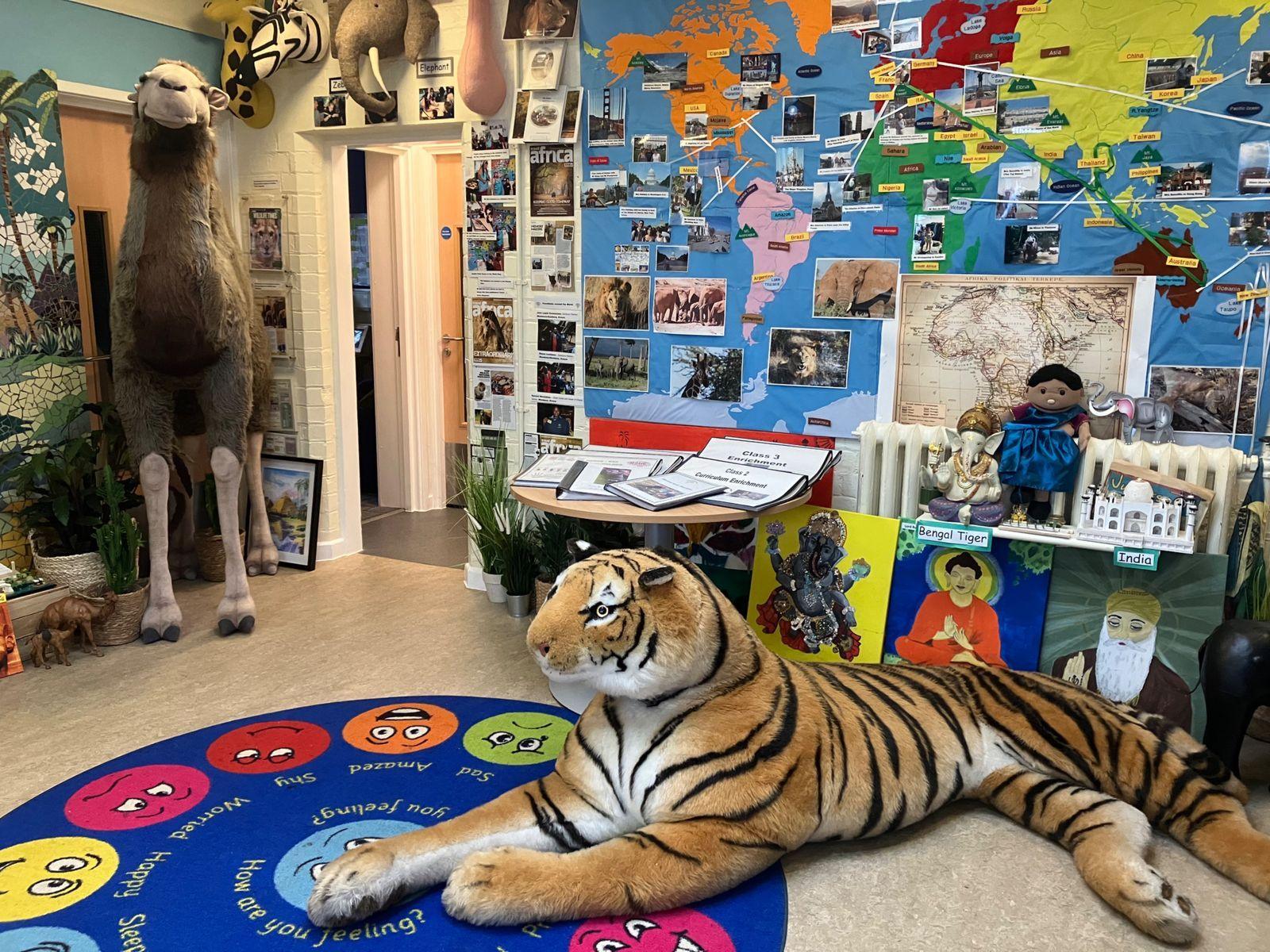 A large stuffed tiger and camel are the main features of a colourful area in the pupil referral unit. The tiger sits on a circular, bright blue mat with lots of different coloured faces on it. There is a map of the world on the wall with lots of images stuck to it.