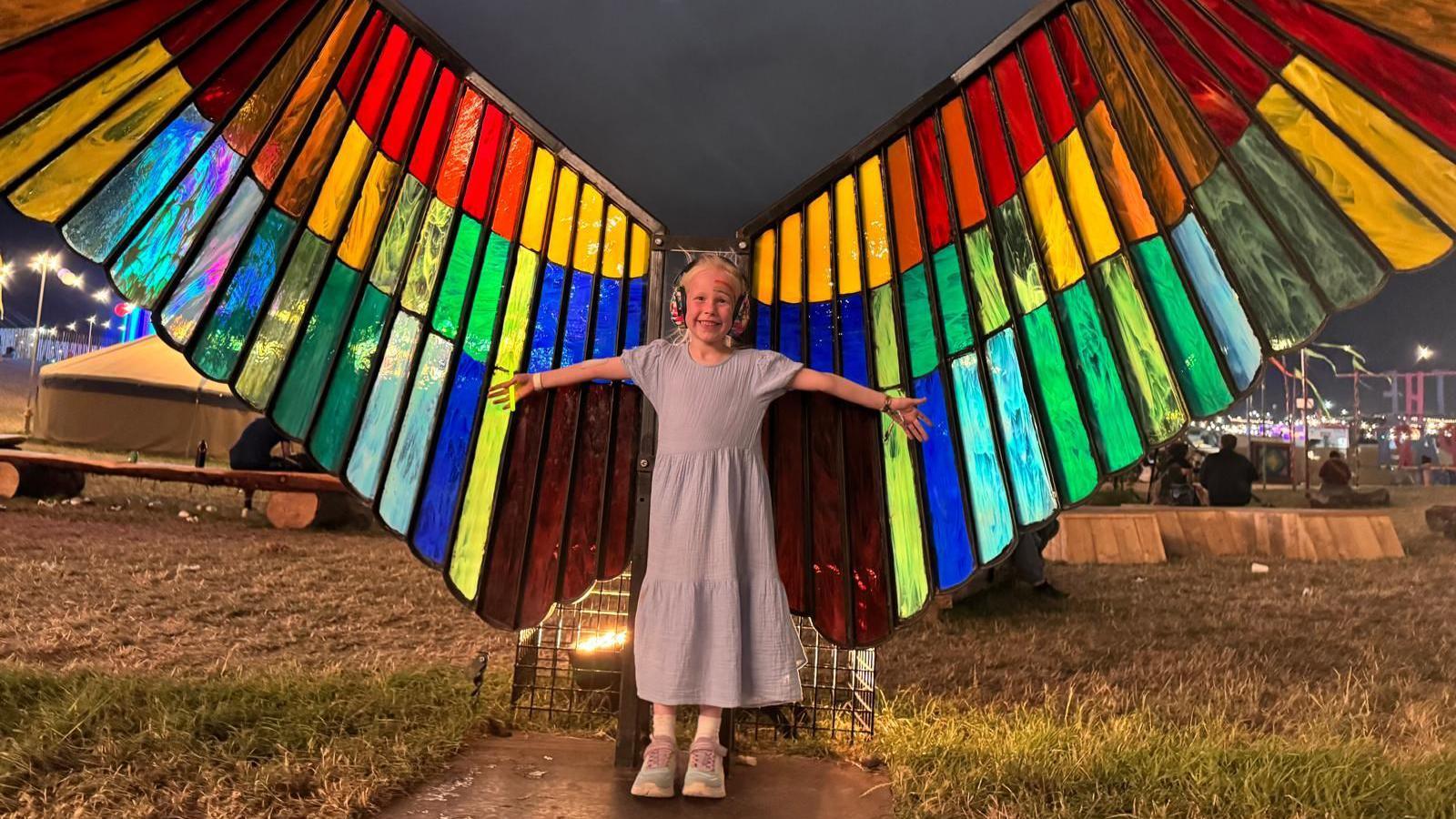 A girl stands with her arms outstretched in front of large, rainbow-coloured angel wings made out of stained glass. The wings are taller than her and span for metres on either side. The girl has ear defenders on and is wearing a blue dress.