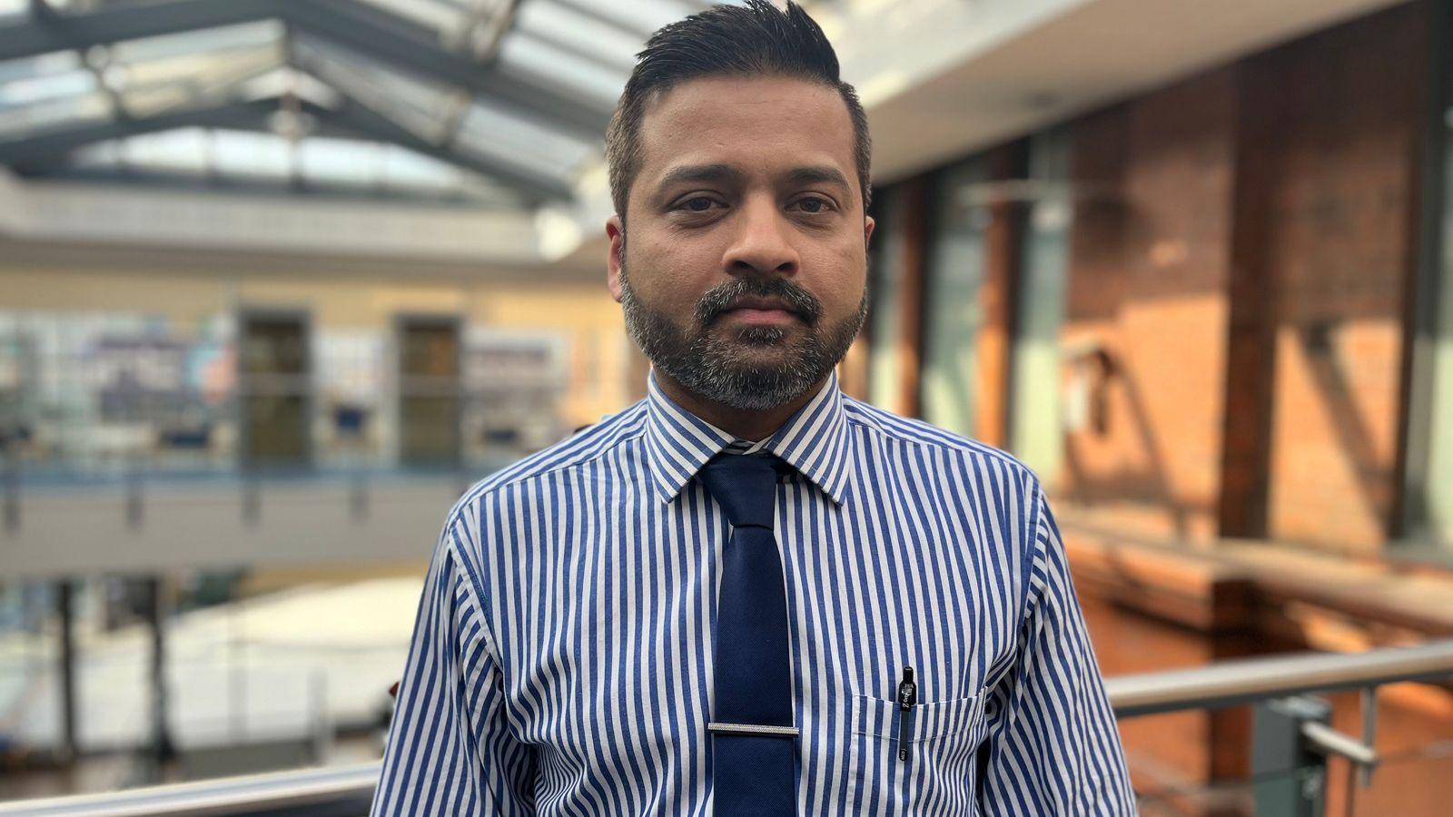 A man with short, black, slightly spiky hair and a black beard flecked with grey. He is standing on a mezzanine with the main atrium of a building blurred in the background. He wears a blue and white vertically striped shirt. A pain dark blue tie is held by a straight tie pin and a pen can be seen poking from his breast pocket.