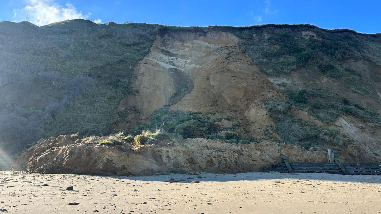 Part of a cliff has fallen away and is on the beach below. Wooden structures can also be seen on the sand.