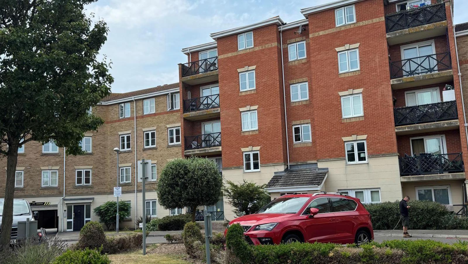 A block of flats, with a red car parked outside beside a small garden