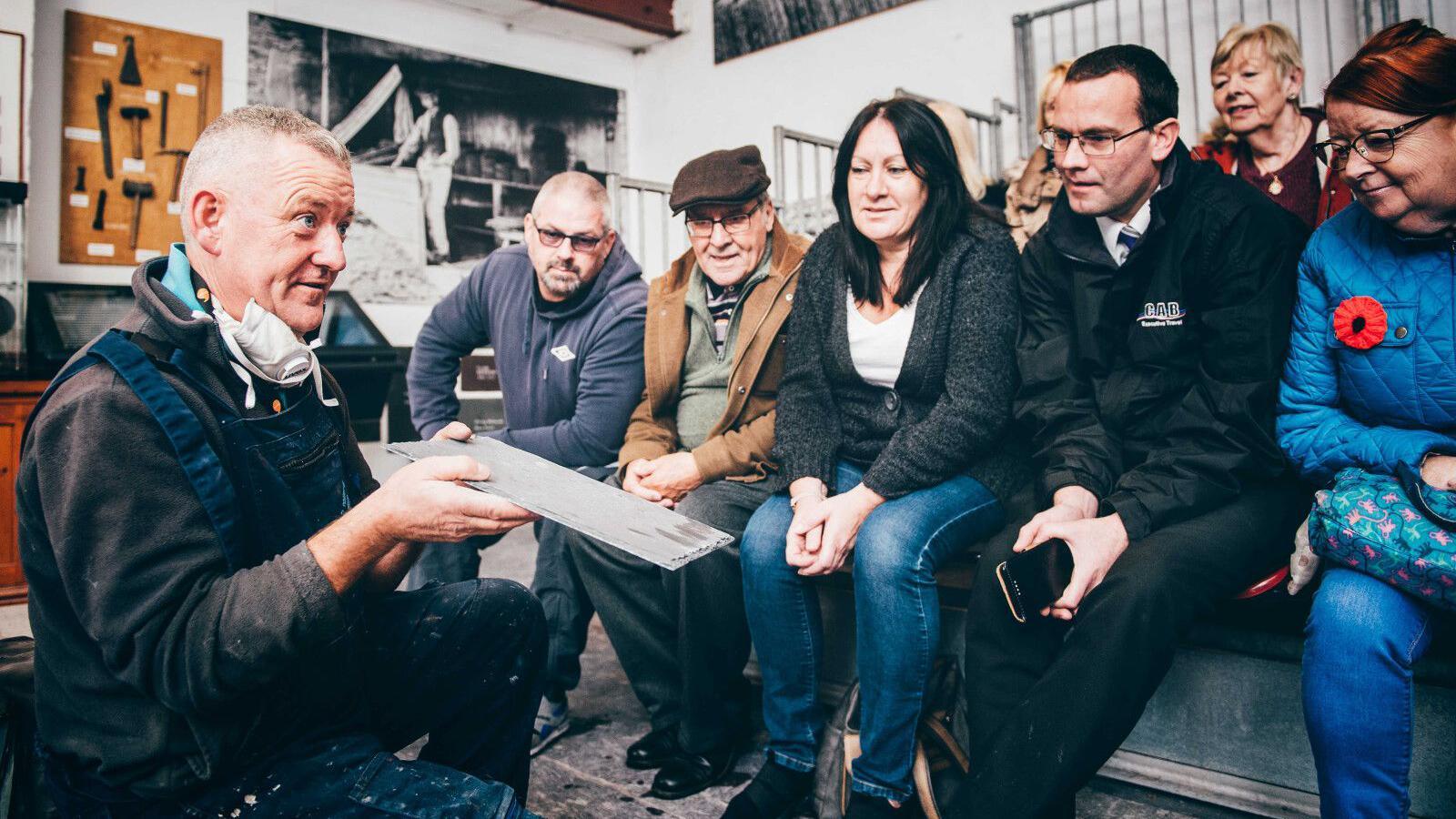 A workman shows an audience sitting nearby a slate that he has just split from the block by hand