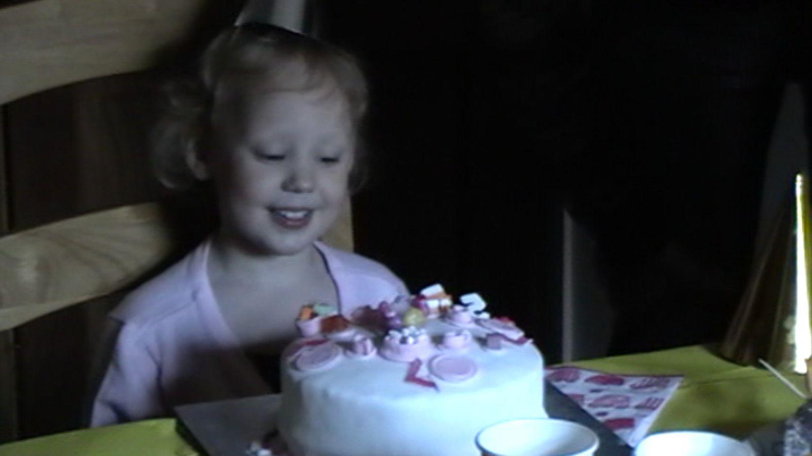 A young girl sat at a table looking down and smiling at a white and pink birthday cake. She has short blonde hair and is wearing a pink top. It looks like it is a screenshot from a home video.