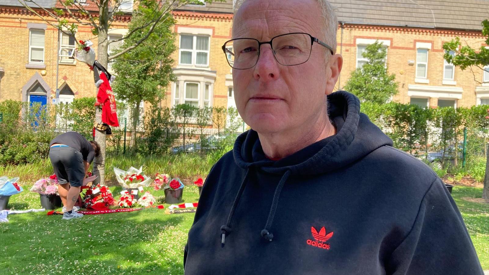 John Barlow, who has light grey hair and is wearing glasses and a dark blue Adidas branded hoodie, looks into the camera with a solemn expression while in the backdrop a man lays flowers by a tree surrounded with tributes. 