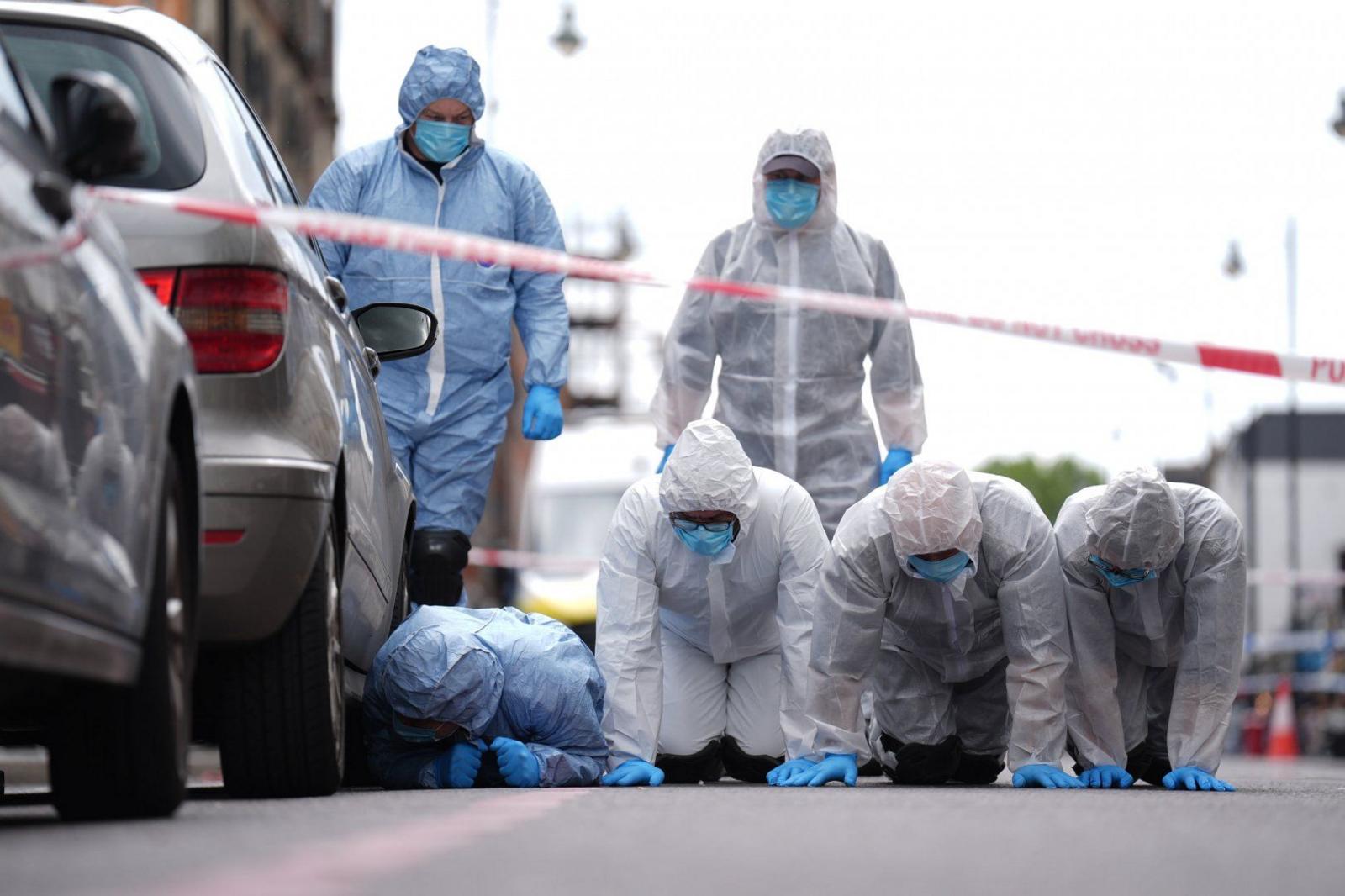 Four people wearing forensic suits, and blue gloves and face masks, are on their hands and knees, searching the street beside the crime scene, with two others standing behind them