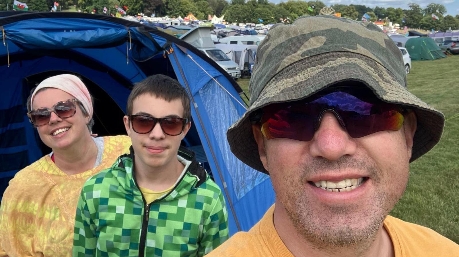 Three people smiling in a field with tents. All of them are wearing sunglasses. Louise is in the back wearing a pink headband and yellow-orange tie dye poncho. Sam has short brown hair and is wearing a checked green hoodie. Chris is wearing a green camo bucket hat and a yellow T-shirt.
