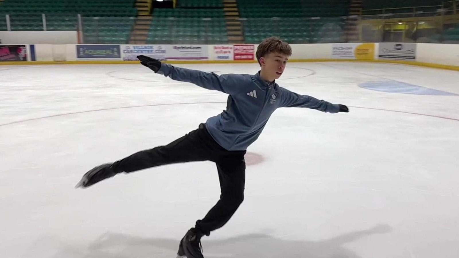 Edward, skating on Whitley Bay Ice Rink, stretches out with one leg high in the air. He's wearing his blue Team GB kit. 