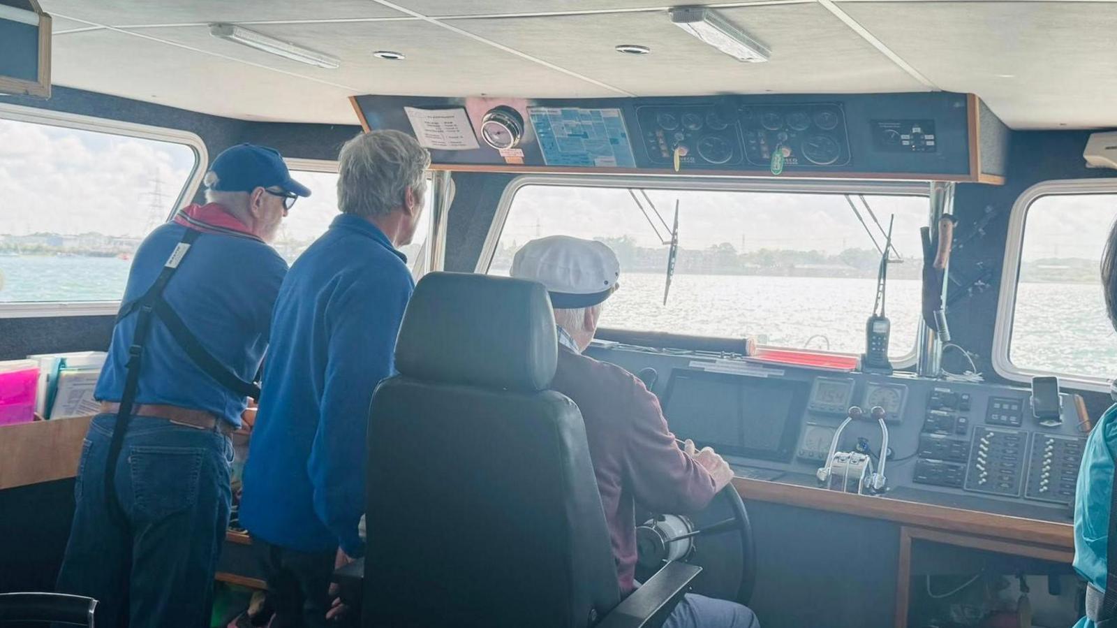 Roger Barnham, wearing a sailor's hat, is sat holding the wheel of a vessel whilst two crew members watch over him.