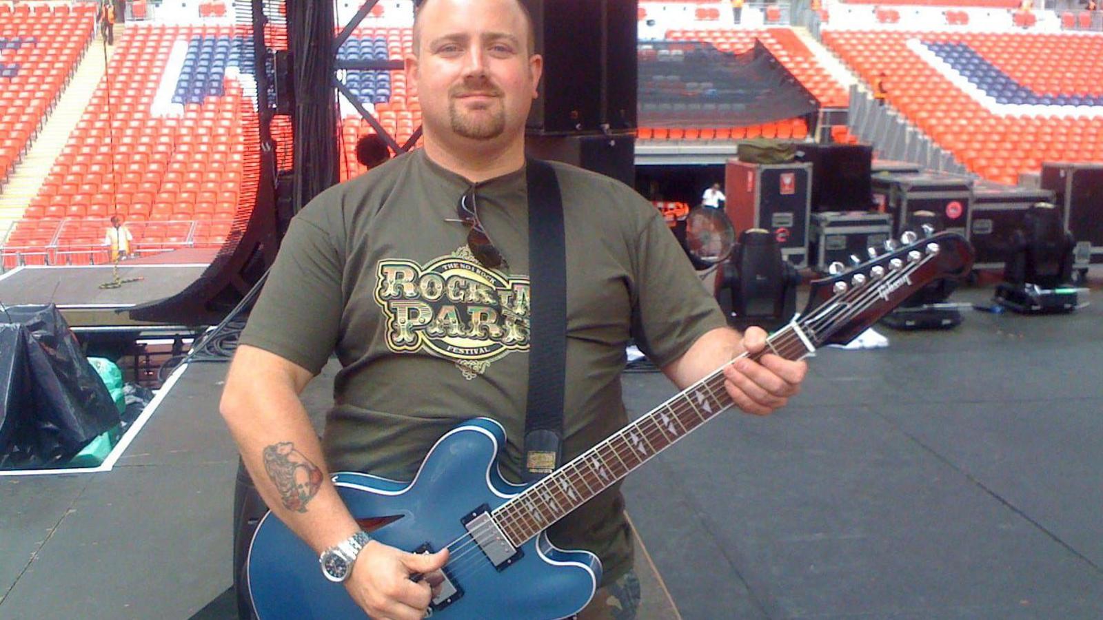 A photo of Alex holding a blue guitar, standing on a stage with rows and rows of red plastic seating visible beyond. Alex is wearing camouflage trousers and a khaki top that reads 'Rocking Party' in gold letters.
