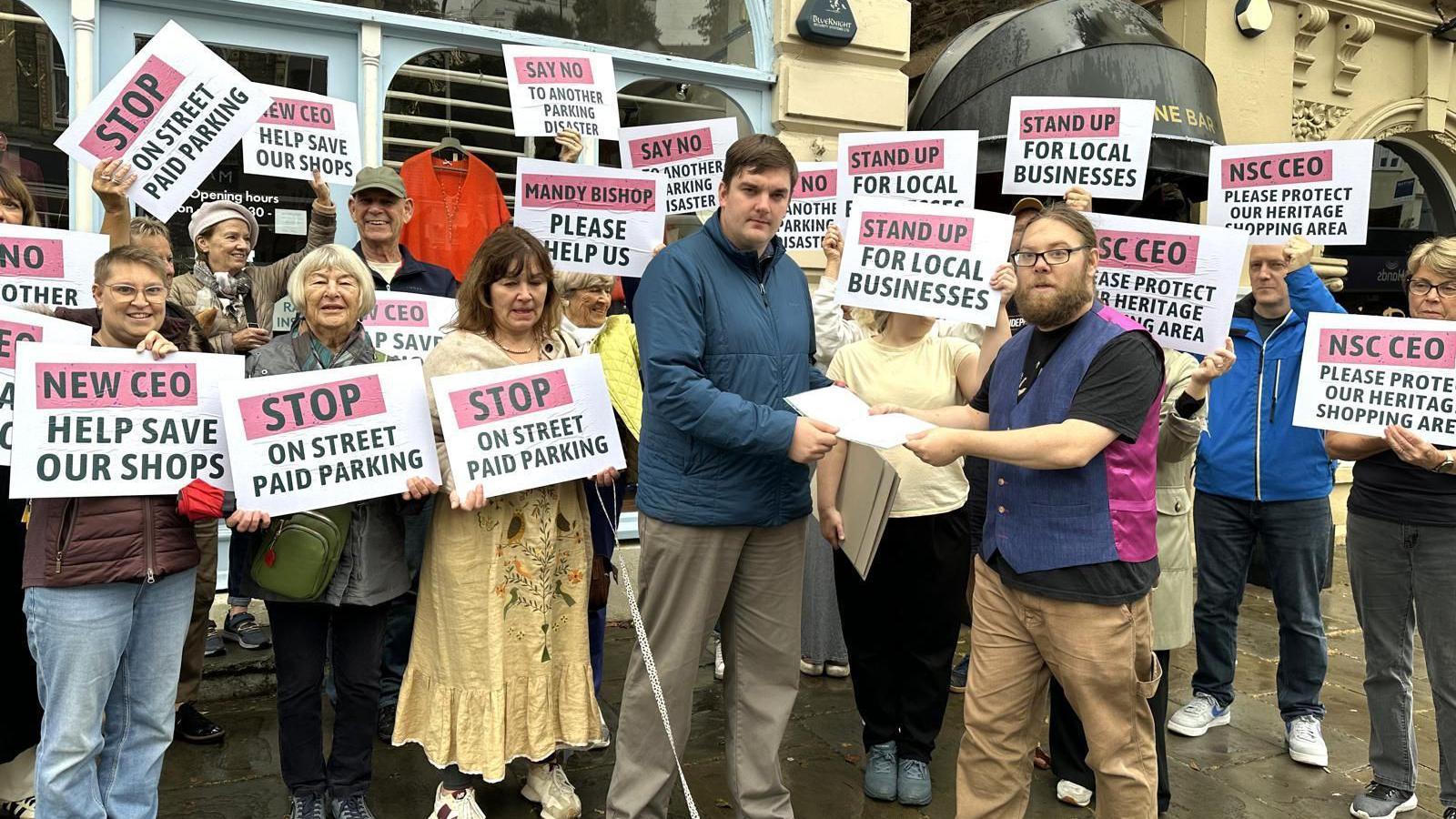 People are holding signs with pleas to stop parking charges. In the foreground, two men are handing over a paper with signatures.