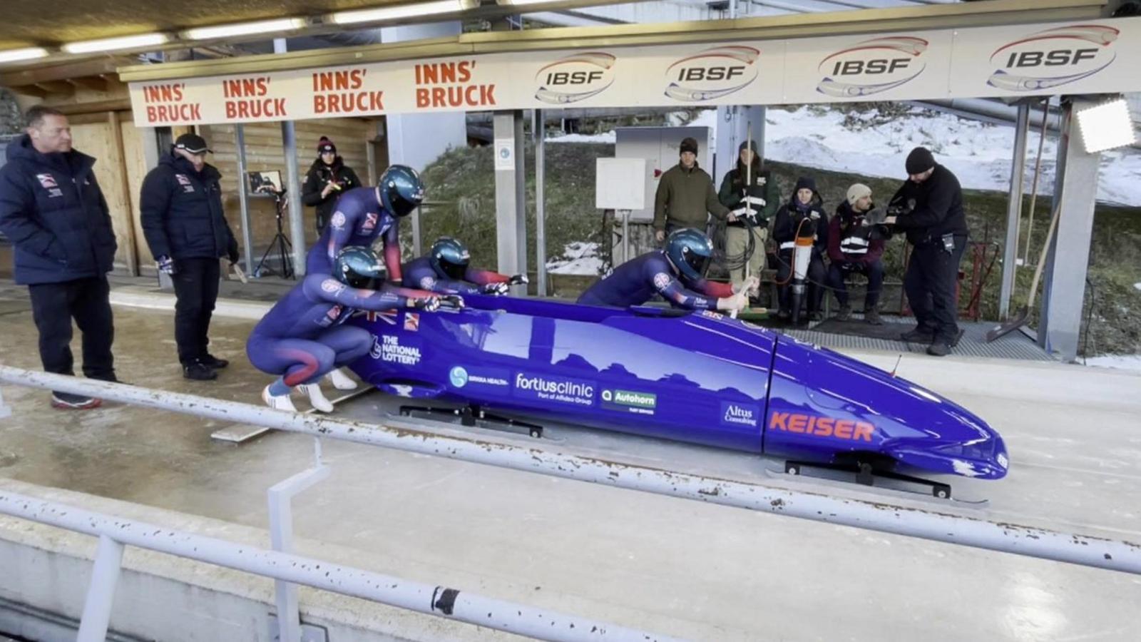 Four men wearing skintight blue race suits are standing next to a blue bobsleigh and preparing the push it down the track. They are being watched by members of the Team GB coaching team.