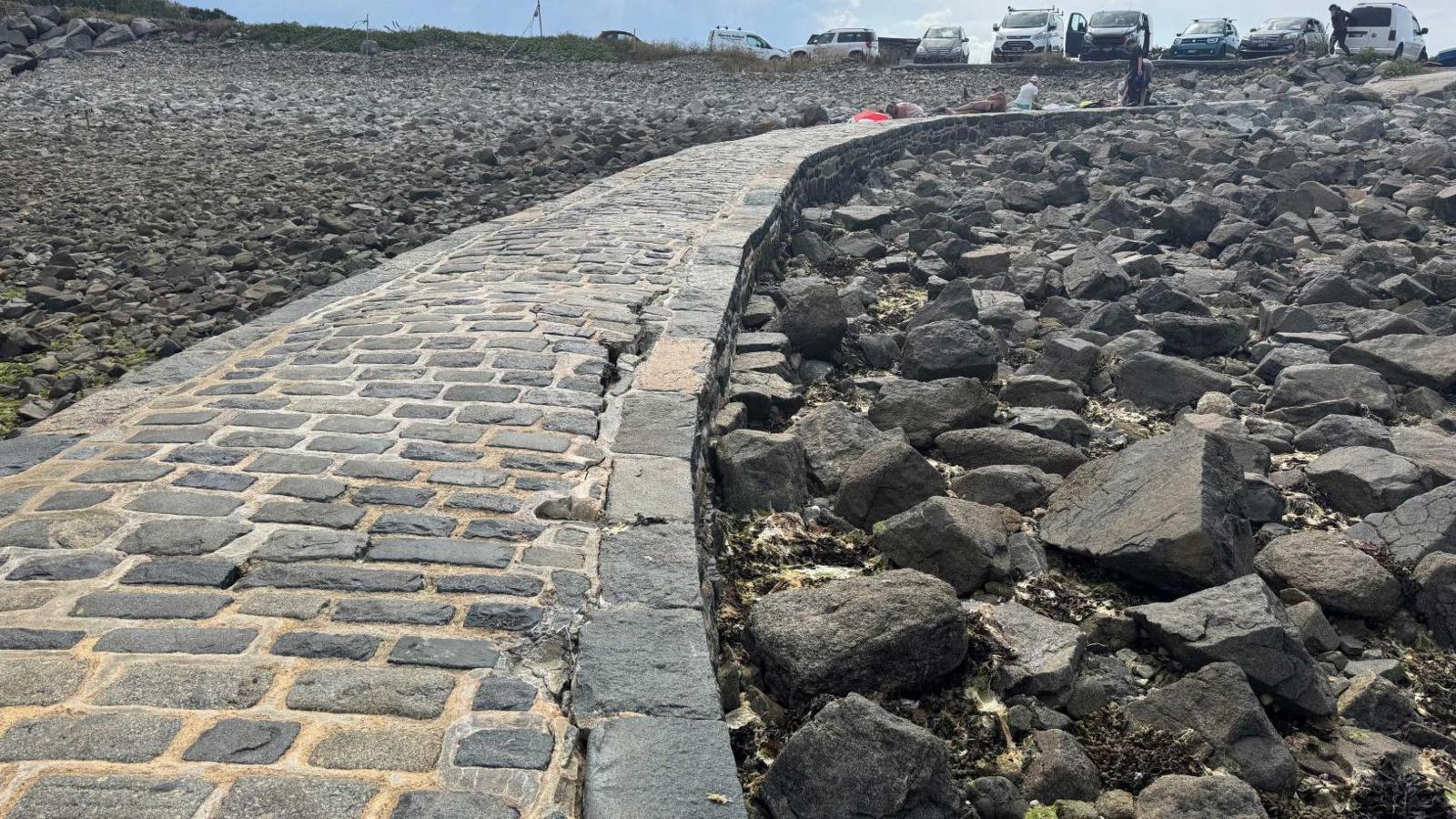 A grey cobblestone pier leading up to a car park. There is a large crack where the pier is raised slightly and bulging.