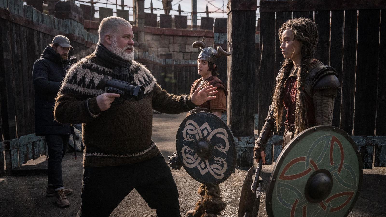 A white haired bearded man gives direction to an actor. He wears a brown and cream nordic knit, holds a camera in his hand and gestures with the other hand. The actor he gestures to is a girl with long brown hair, dressed in viking warrior clothing and she holds a round colourful shield