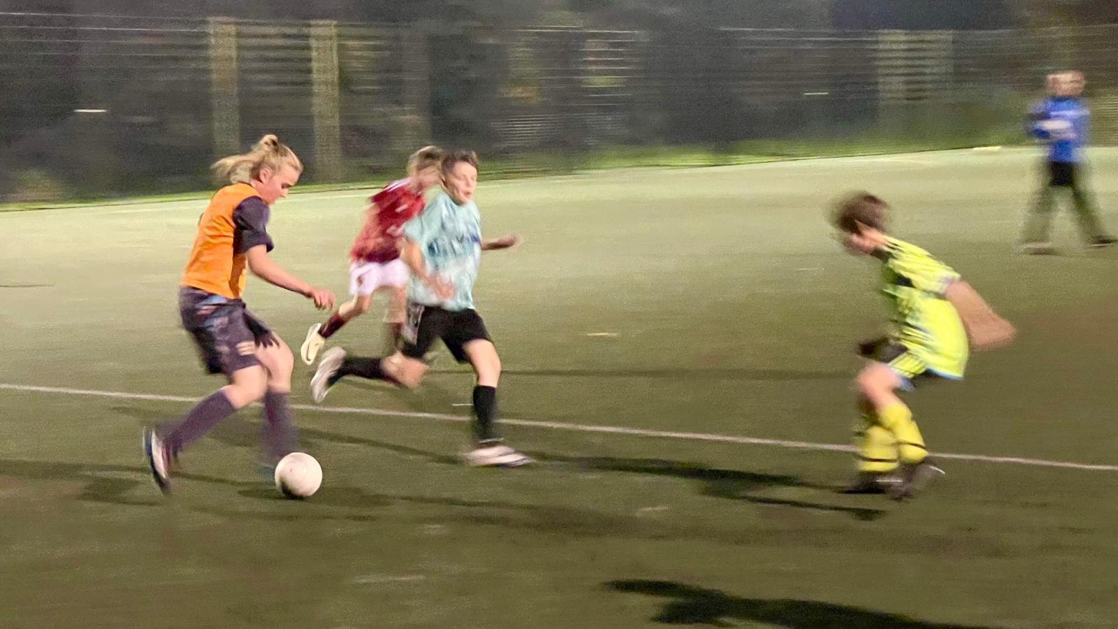 A girl with blonde hair tied back in a tail is running towards three boys with a football at her feet on an astro turf. She is wearing a dark purple England football kit and an orange bib. One of the boys is wearing a red Manchester United kit with white shorts, another is wearing a green and white t-shirt with black shorts and socks and another boy is wearing a full lime green Arsenal kit. A man in a blue tracksuit top and black bottoms can be seen in the background. 