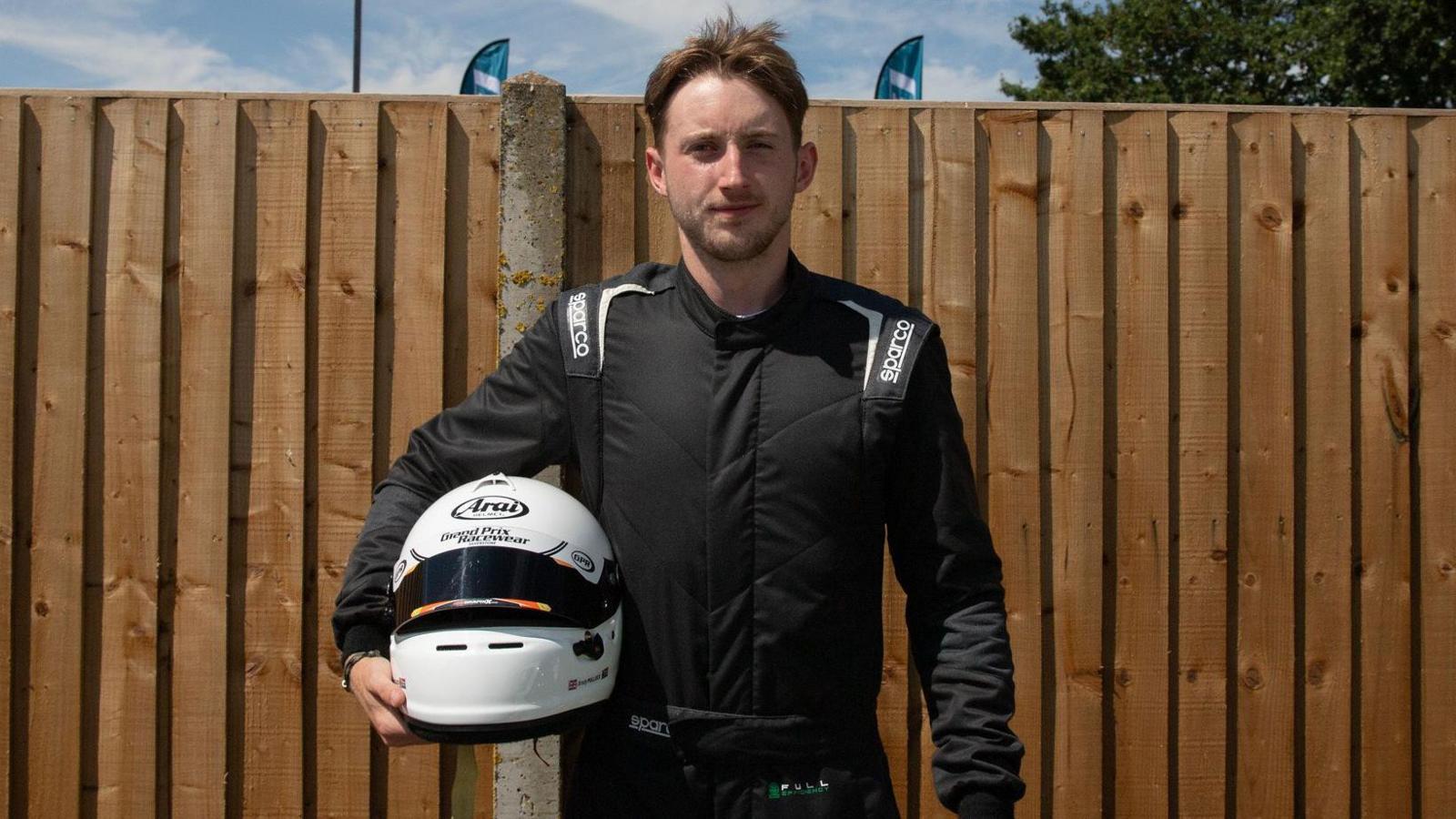 Brady Pollock standing in front of a brown fence, dressed in all-black race gear whilst holding his white crash helmet.