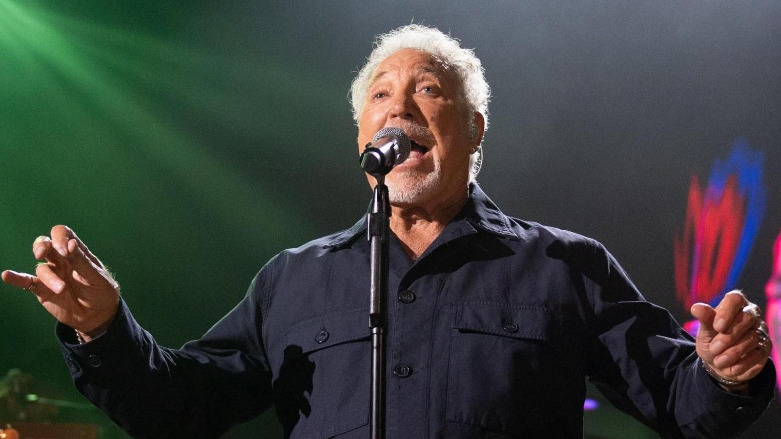 Sir Tom Jones on stage singing into a microphone. He is wearing a black shirt and has his arms stretched.