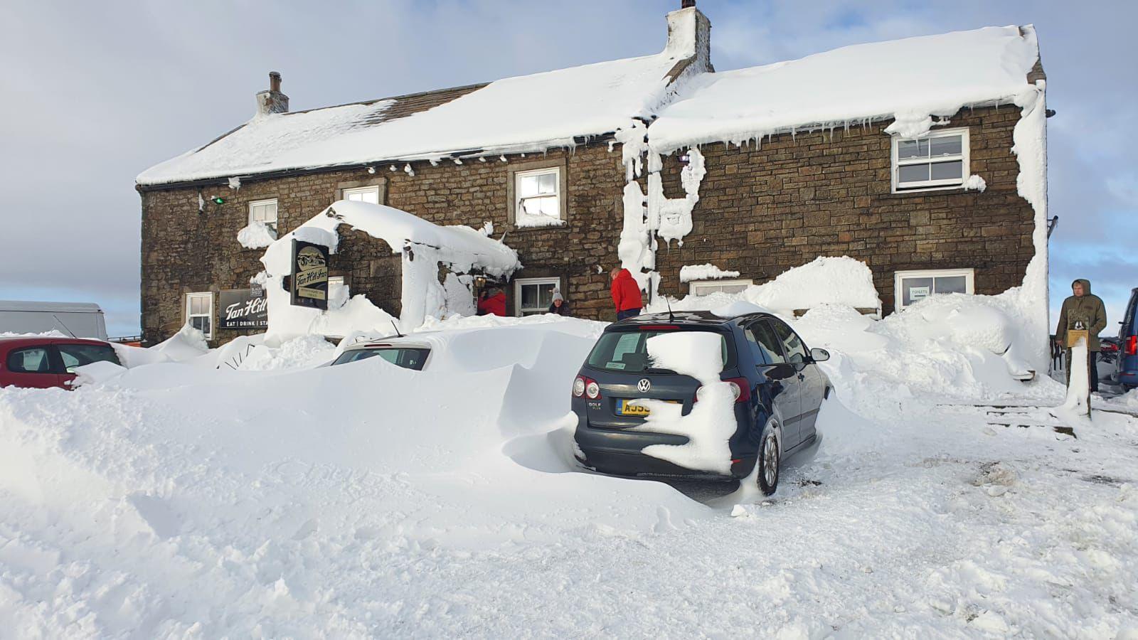 Snow on Tan Hill Inn in the Yorkshire Dales on November 27th 2021. A beige stone building is covered in snow, with several cars parked outside also buried under thick snow.