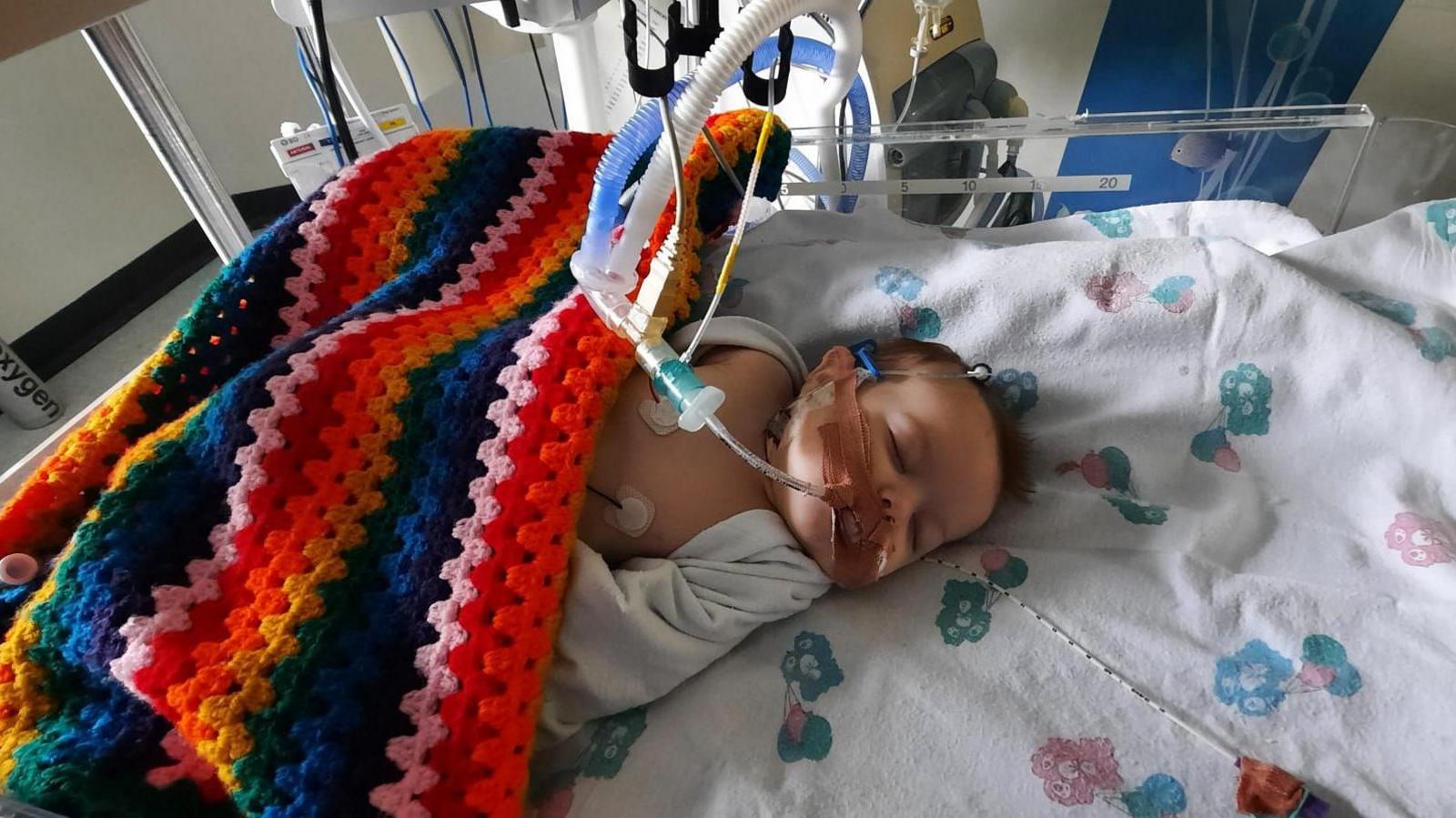 Freya Dixon as a baby in a hospital bed covered in a brightly coloured blanket with tubes in her mouth