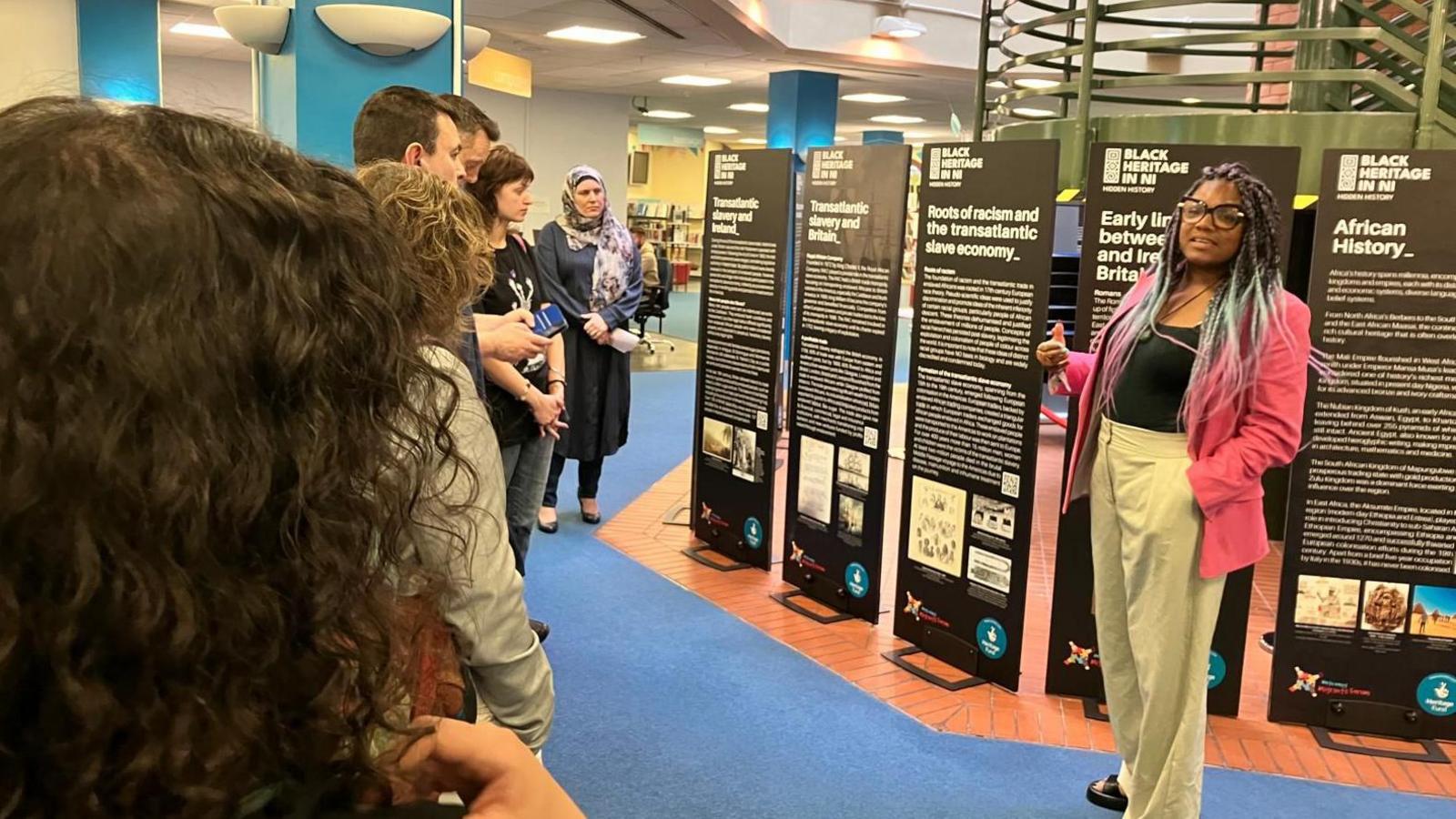 Chantelle Lunt is giving a talk at the opening of the museum exhibit. 
A crowd of people are stood listening to her. 
She is wearing a black top, pink jacket and cream trousers. 