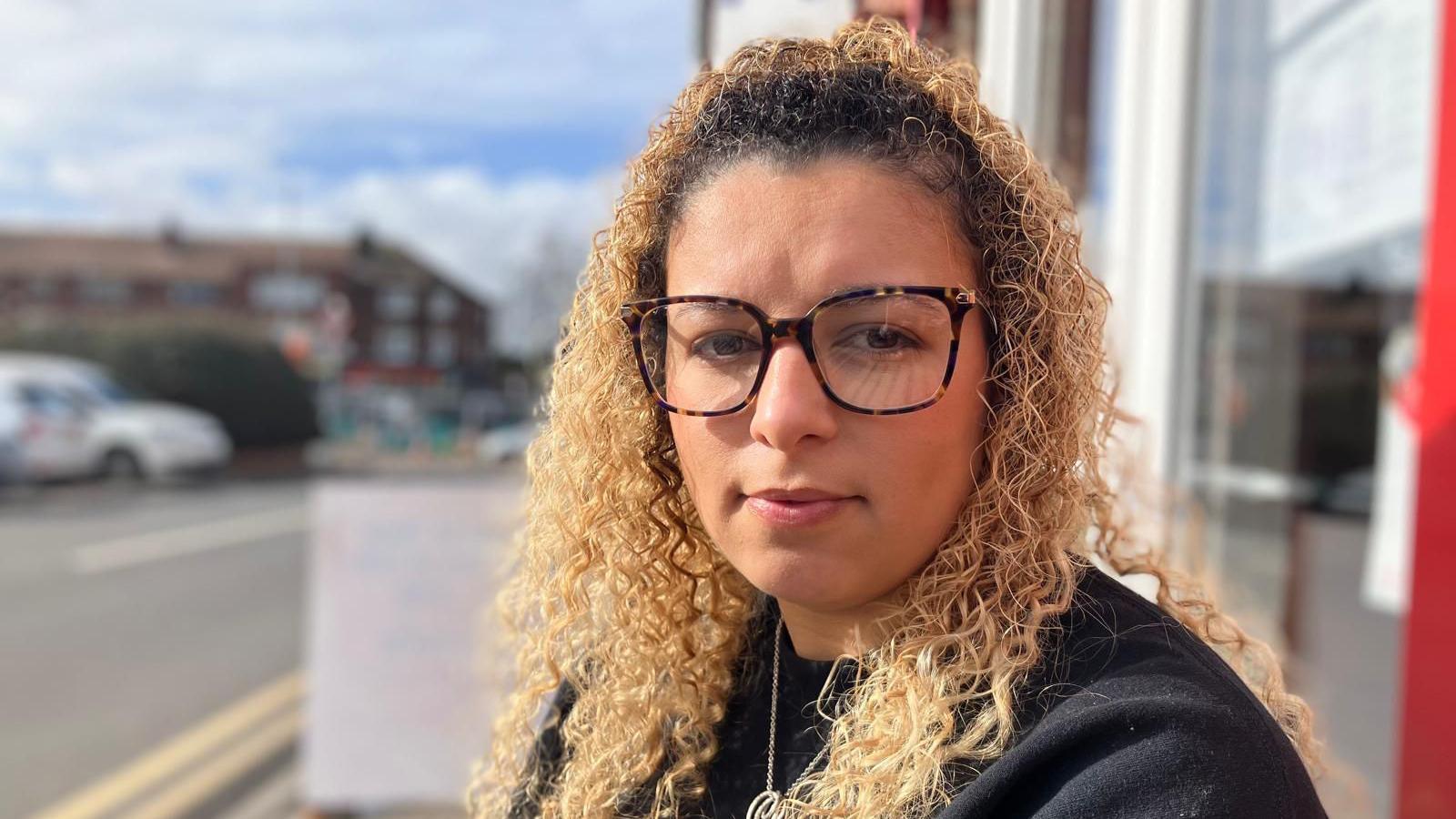 A woman with blonde curly hair and square glasses sits outside her coffee shop. Roadworks in the background are blurred.