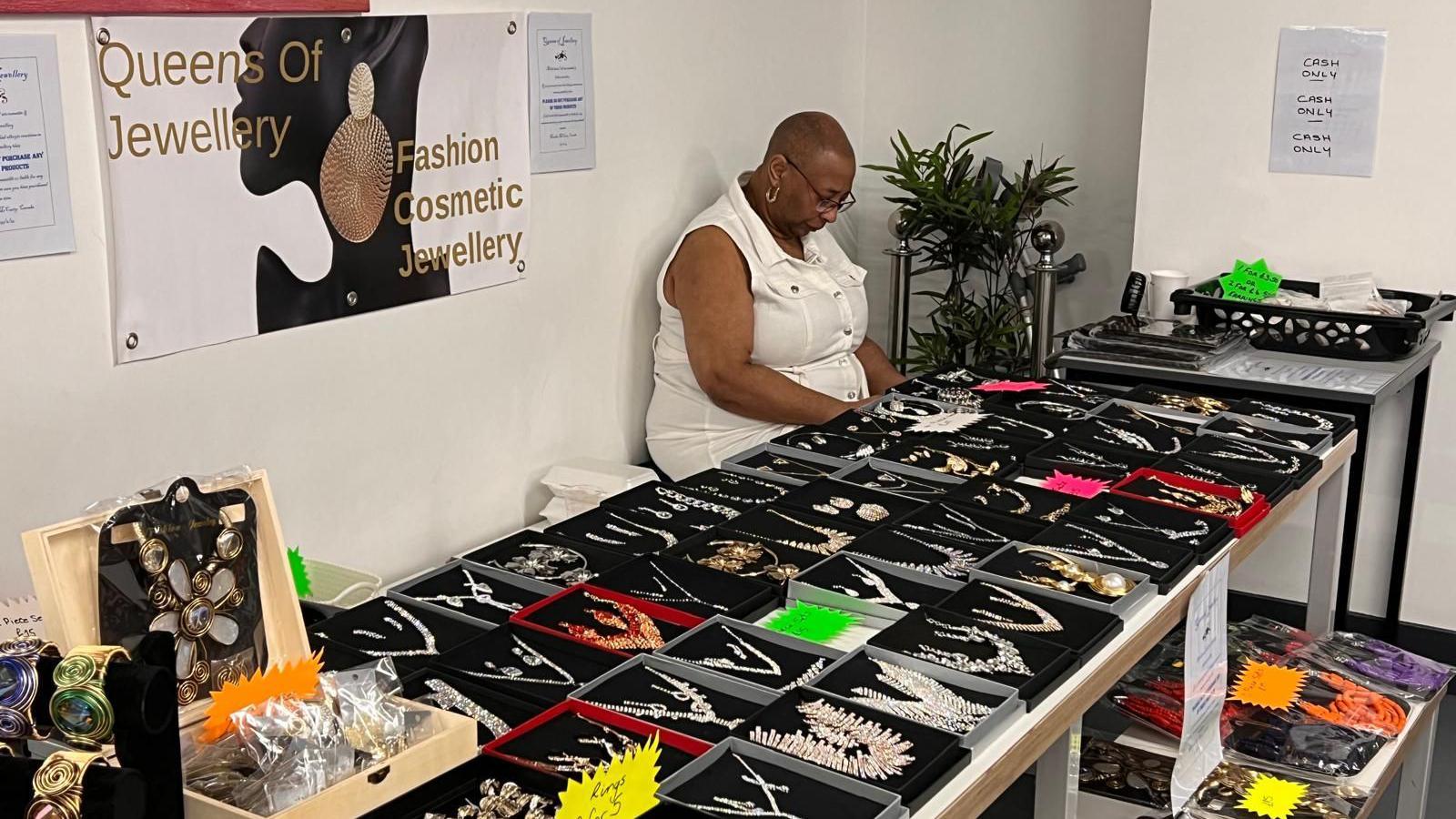 Marsha sits behind a tables which display her jewellery collection. There is a banner on the wall behind her which says 'Queens of Jewellery Fashion Cosmetic Jewellery."